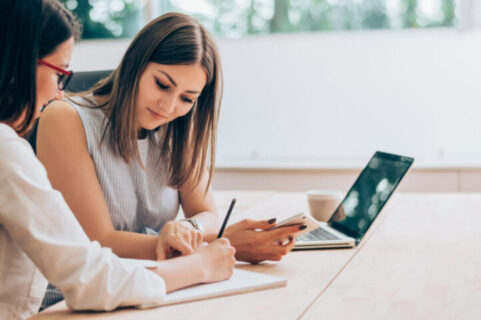 Two female colleagues in an office with an open laptop in front of one whilst the other writes on a notepad with a pen.