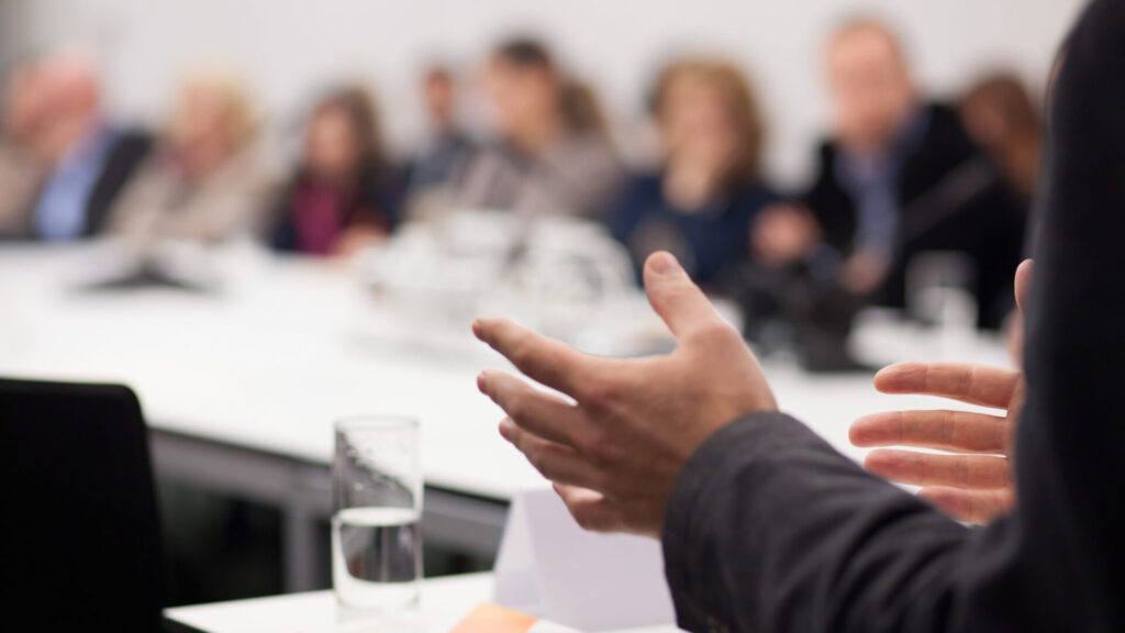A business person chairs a meeting regarding collective redundancies to start collective consultation, with employees blurred in the background in a large corporate meeting room.