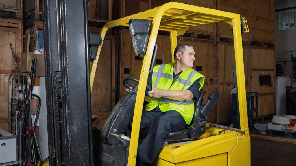 Man wearing high visibility vest reversing forklift truck inside warehouse while looking behind.