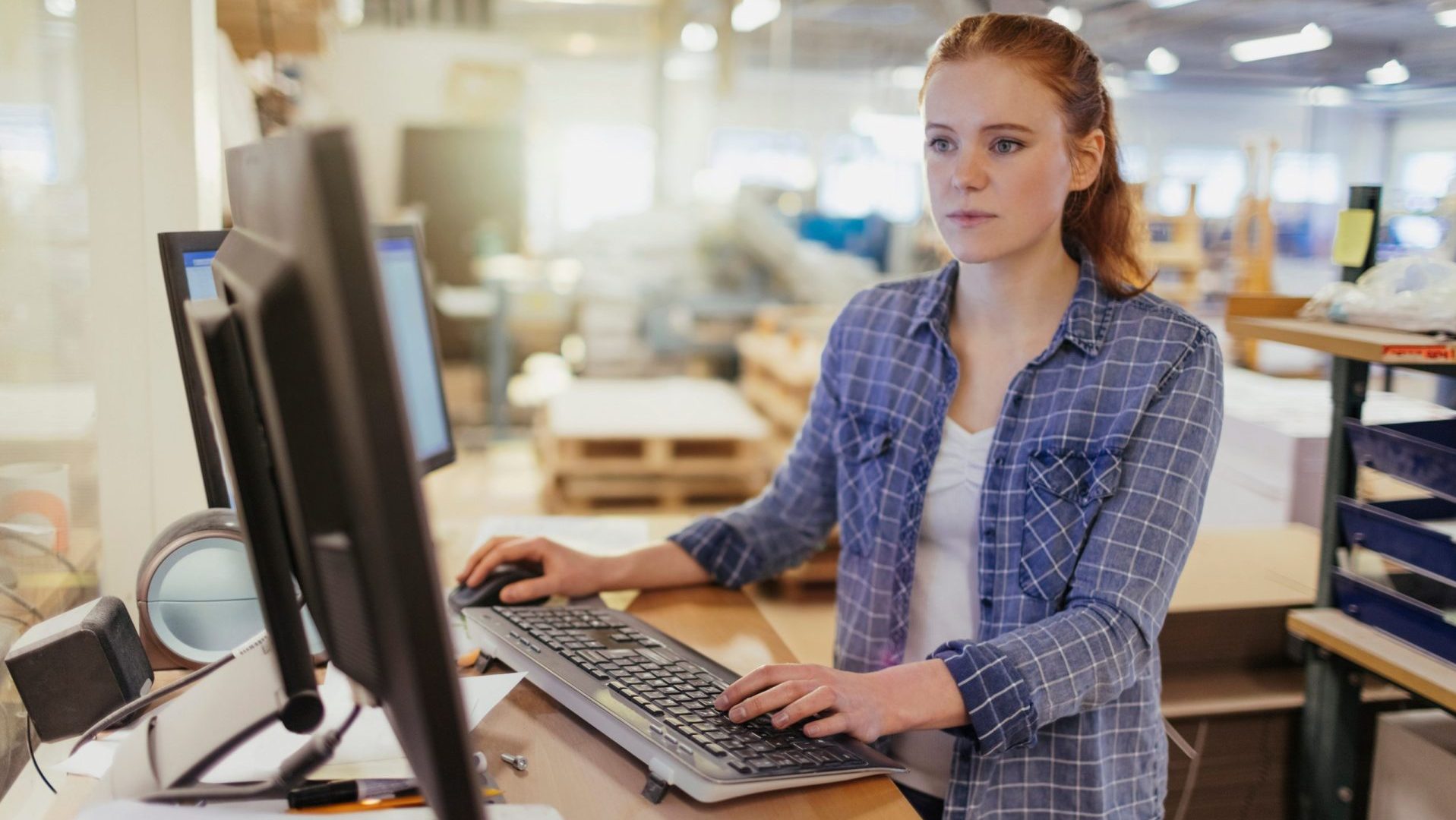 An office worker in casual attire works on two monitors at a standing desk with a warehouse environment in the background.