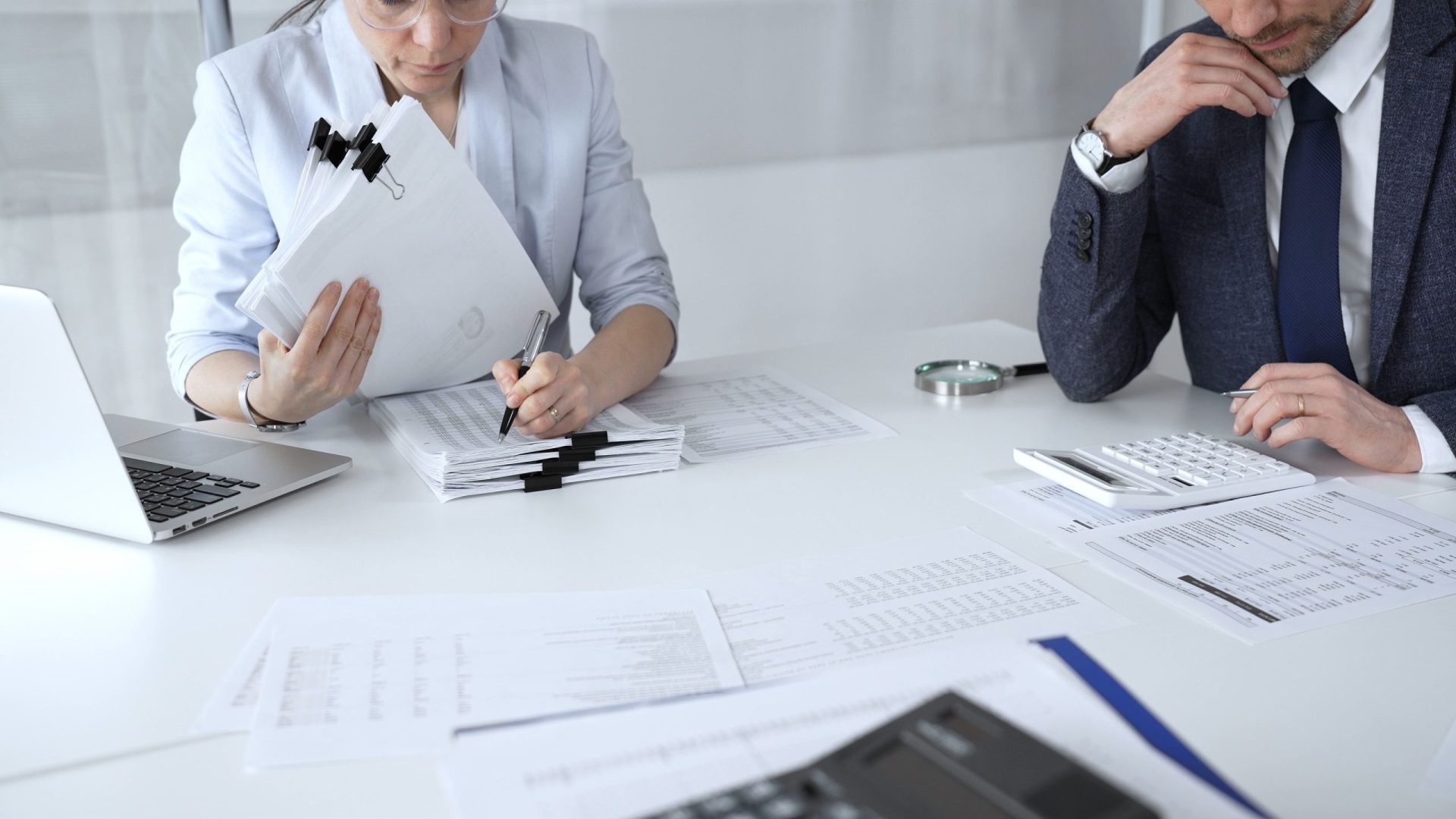 Two business people sit at a desk in front of many documents and a laptop to carry out internal audits.