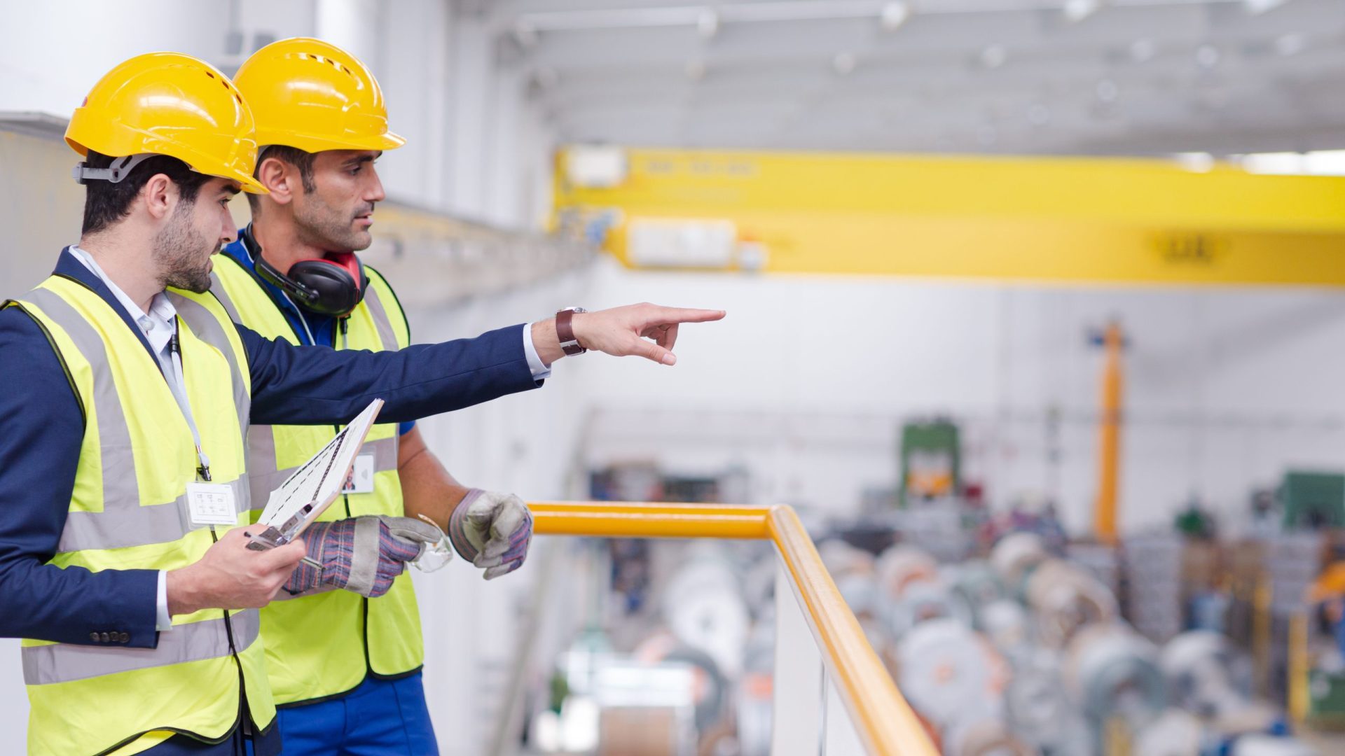 Two safety offices stand on a balcony overlooking a warehouse wearing hard hats and high visibility jackets. One holds a clipboard and is pointing to the distance.