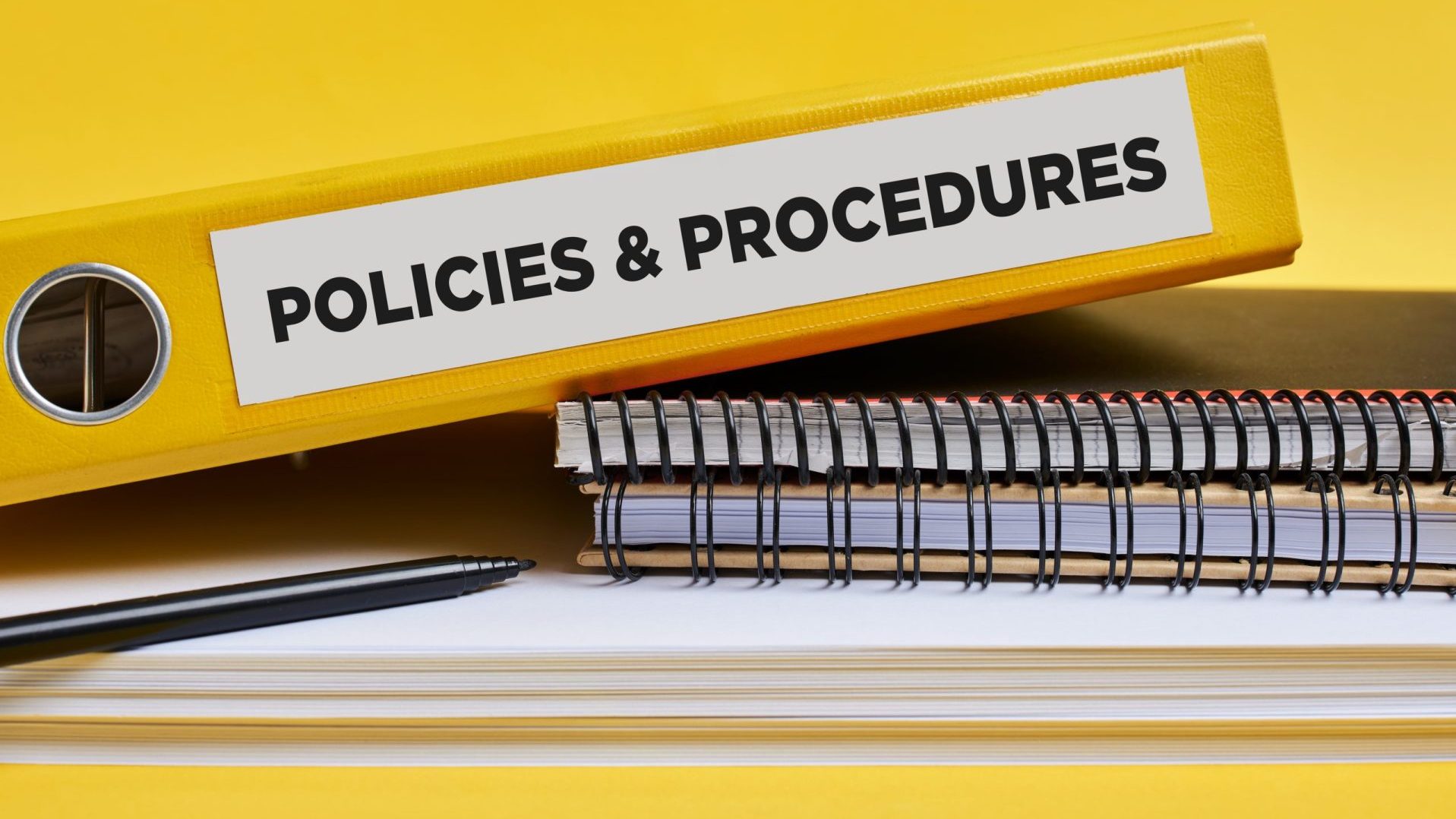 Policies and procedures written on the label of a yellow binder on the office desk.