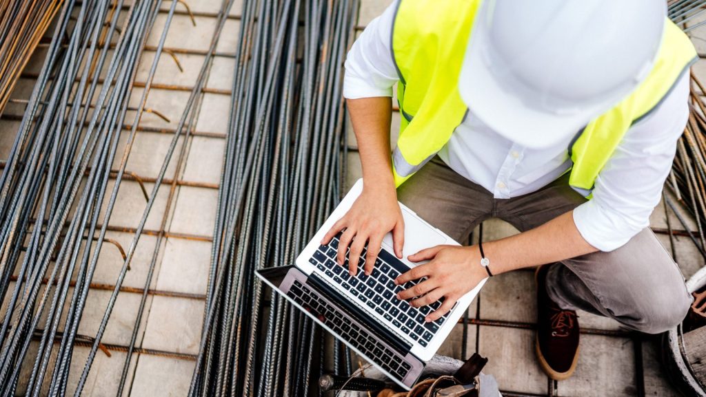A health and safety consultant in high visibility vest and hard hat checks their laptop on a construction site